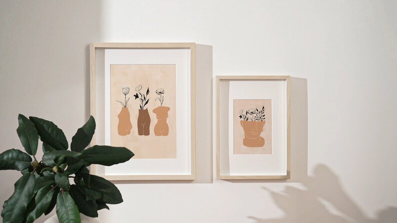 Pu&ograve; includere: Due stampe incorniciate con disegni minimalisti di figure stilizzate con fiori che crescono dalle loro teste e corpi. Le stampe sono su una parete bianca con una pianta verde in primo piano.