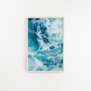 May include: A framed art print featuring an abstract image of ocean waves. The artwork showcases a dynamic interplay of turquoise and white, capturing the movement and texture of the water. The print is encased in a light-colored wooden frame.