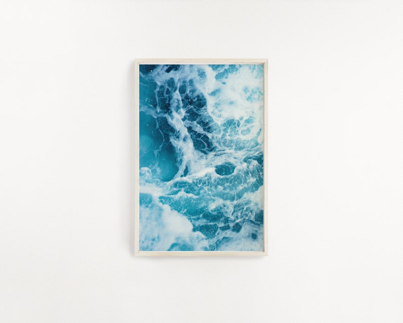 May include: A framed art print featuring an abstract image of ocean waves. The artwork showcases a dynamic interplay of turquoise and white, capturing the movement and texture of the water. The print is encased in a light-colored wooden frame.