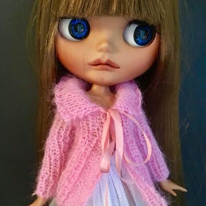 Op de afbeelding: Een pop met lang bruin haar, die een roze gebreide trui draagt met een witte kanten rok en blauwe ogen.