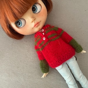 May include: A doll with red hair and blue eyes wearing a red and green sweater and light blue jeans. The doll's sweater has green stripes and white buttons. The doll is lying on a gray surface.