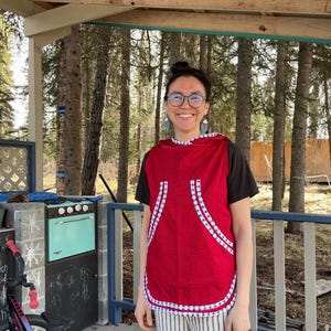 May include: A red hooded vest with white and blue patterned trim and accents. The vest is worn over a black t-shirt and paired with striped trousers. The background includes a bicycle, a vintage teal oven, and a wooden porch.