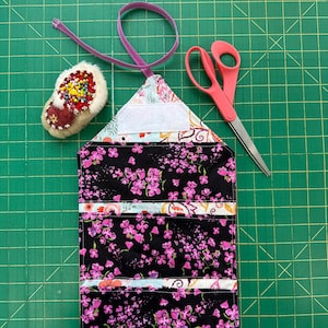 May include: A handmade sewing kit with a black floral print, pink scissors, and a pincushion. The kit has multiple pockets and a purple elastic closure. The pincushion is red and white with colorful pins.