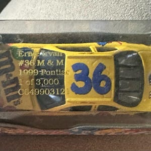 Puede incluir: Un coche amarillo de fundición a presión con el número 36 en el lateral y las palabras "M&M's" en el lateral. El coche está en una caja de plástico transparente con un respaldo de cartón azul y blanco. El respaldo tiene las palabras "Action" y "NASCAR" en él.