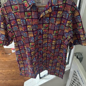 May include: A purple, red, yellow, and blue short-sleeved button-down shirt with a repeating pattern of the text "Imagine" and "Free" and images of a roller coaster, a spaceship, and a sphere. The shirt has a tag that reads "Disney Parks Merchandise."