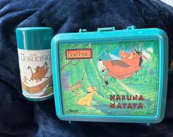 Może przedstawiać: Turkusowa niebieska lunchbox z motywem Króla Lwa. Przód lunchbox przedstawia scenę z filmu z Pumbaą, Timonem i Simbą. Na lunchboxie nadrukowany jest tekst "Hakuna Matata". Lunchbox została wyprodukowana przez Aladdin.