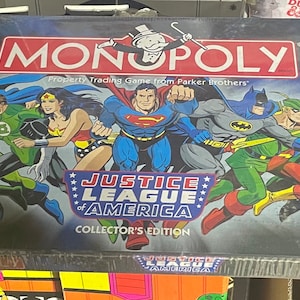 May include: A Monopoly board game box featuring the Justice League of America. The box is black with a silver Monopoly logo and a Justice League of America logo. The box features images of Superman, Batman, Wonder Woman, Green Lantern, Aquaman, The Flash, and Green Arrow.