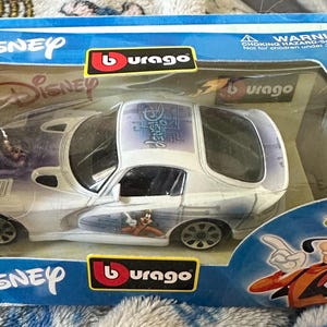 May include: A Disney-themed toy car in a blue and clear plastic box. The white car has purple and blue graphics and features Disney characters. The box has the Disney and Burago logos, and a warning for children under 3 years.