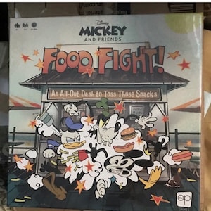 May include: A board game box titled "Mickey and Friends Food Fight!" features Disney characters in a food fight scene. The box art includes the game title, character illustrations, and the tagline "An All-Out Dash to Toss Those Snacks."