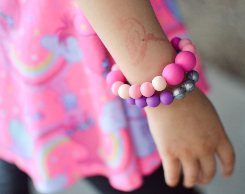 May include: Two beaded bracelets in shades of pink, purple, and silver. The bracelets are made of round beads and are worn on a person's arm. The background is a pink dress with a unicorn and rainbow design.