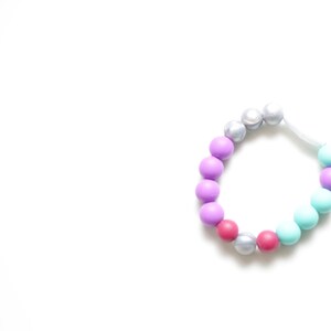 May include: A multi-coloured beaded bracelet. The beads are in shades of purple, pink, silver, and teal. The bracelet is strung on a white elastic cord. The bracelet is set against a white background.