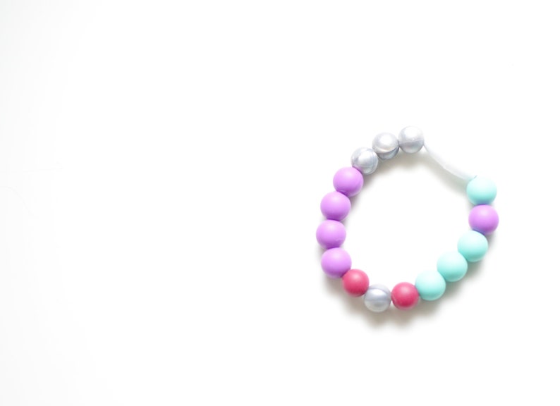 May include: A multi-coloured beaded bracelet. The beads are in shades of purple, pink, silver, and teal. The bracelet is strung on a white elastic cord. The bracelet is set against a white background.