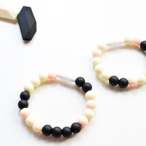 May include: Two beaded bracelets in black, cream, and pink. The bracelets are made of round beads and are connected by a white, cylindrical piece. A black and white geometric shape is in the upper left corner.