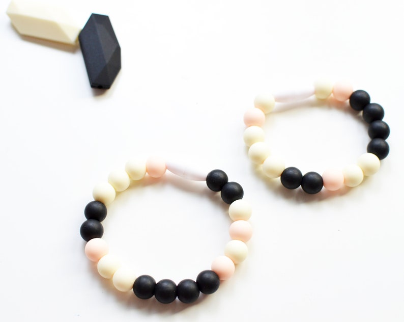 May include: Two beaded bracelets in black, cream, and pink. The bracelets are made of round beads and are connected by a white, cylindrical piece. A black and white geometric shape is in the upper left corner.