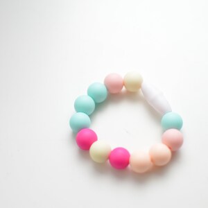 May include: A pastel-coloured bracelet featuring a series of round beads in shades of pink, blue, and yellow. The beads are strung together with a white clasp. The bracelet is designed for babies and toddlers.