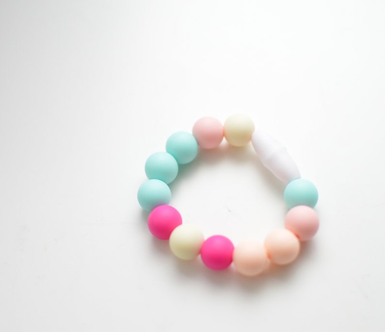 May include: A pastel-coloured bracelet featuring a series of round beads in shades of pink, blue, and yellow. The beads are strung together with a white clasp. The bracelet is designed for babies and toddlers.