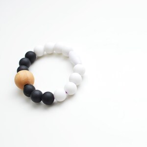 May include: A bracelet made of black, white, and a single wooden bead. The bracelet has a gradient effect, transitioning from black to white beads. The wooden bead provides a natural contrast. The beads are likely silicone or plastic.