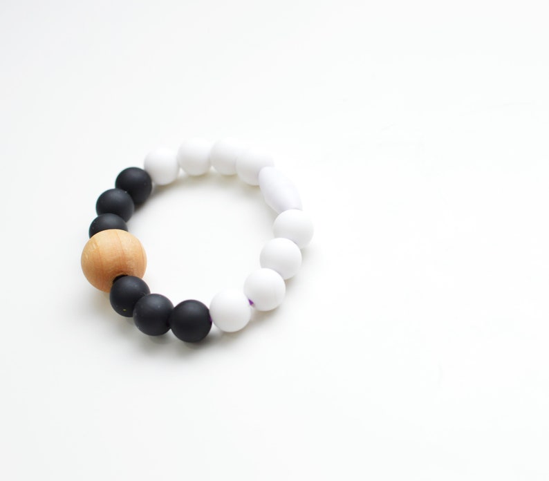 May include: A bracelet made of black, white, and a single wooden bead. The bracelet has a gradient effect, transitioning from black to white beads. The wooden bead provides a natural contrast. The beads are likely silicone or plastic.