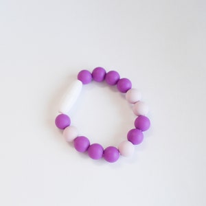 May include: A bracelet made of round beads in shades of purple and light pink, with a white clasp. The beads are arranged in an alternating pattern. The bracelet is a fashion accessory and is displayed on a white surface.
