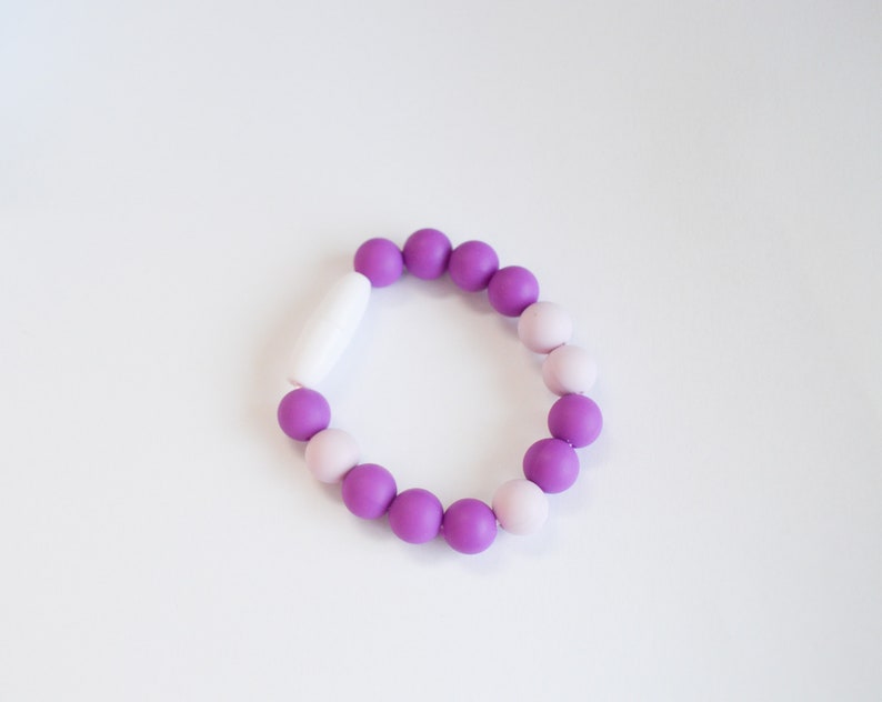 May include: A bracelet made of round beads in shades of purple and light pink, with a white clasp. The beads are arranged in an alternating pattern. The bracelet is a fashion accessory and is displayed on a white surface.