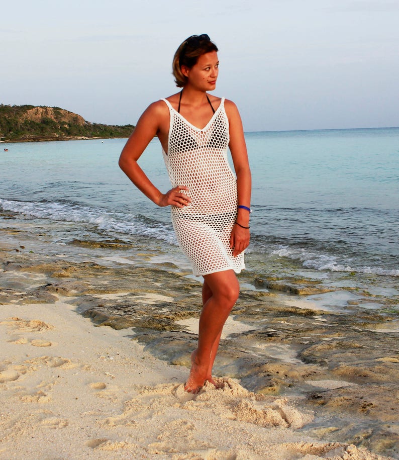 Puede incluir: Una mujer con un vestido de playa de crochet blanco en una playa de arena. El vestido es holgado, sin mangas y tiene un escote en V.