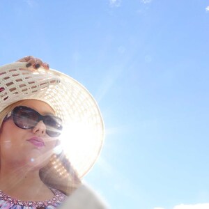 May include: A woman wearing a white crochet sun hat with black sunglasses, looking up at the bright blue sky.