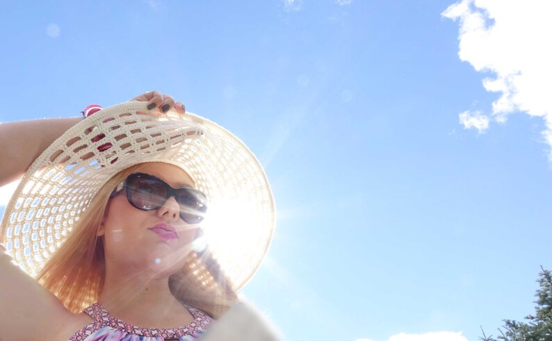May include: A woman wearing a white crochet sun hat with black sunglasses, looking up at the bright blue sky.