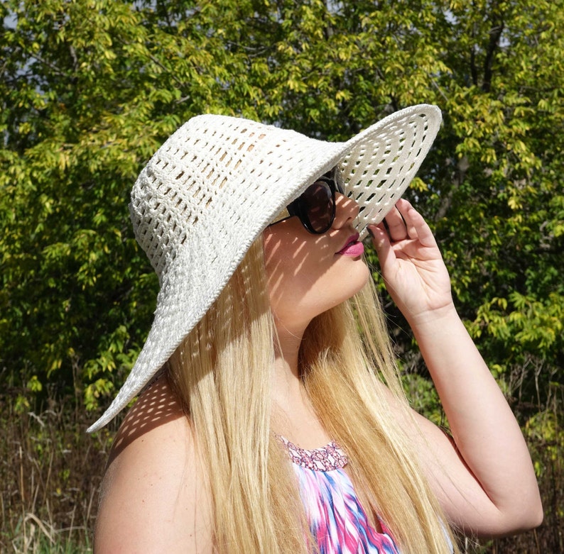May include: A woman wearing a white crochet sun hat with a wide brim and sunglasses. She is looking up at the sun.