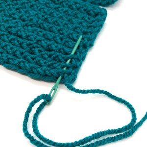 Puede incluir: Primer plano de tejido de punto de color verde azulado con una aguja verde e hilo a juego. La aguja se inserta a trav&eacute;s de la tela, con el hilo enhebrado a trav&eacute;s de su ojo. La textura del material de punto es visible.