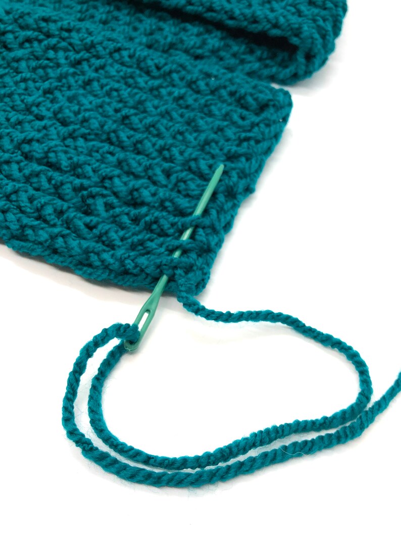 Puede incluir: Primer plano de tejido de punto de color verde azulado con una aguja verde e hilo a juego. La aguja se inserta a trav&eacute;s de la tela, con el hilo enhebrado a trav&eacute;s de su ojo. La textura del material de punto es visible.