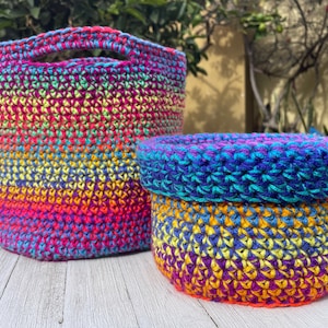 May include: Two handmade crochet baskets in vibrant rainbow colors. The larger basket is cylindrical with a handle, while the smaller one is round with a lid. The baskets feature a mix of pink, yellow, blue, and purple hues, perfect for storage or decor.