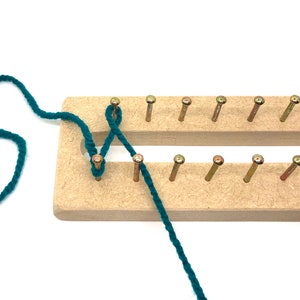 Puede incluir: Un telar de punto de madera con clavijas met&aacute;licas, parcialmente ensartado con hilo verde azulado. El telar es rectangular con dos filas de clavijas. El hilo est&aacute; enrollado alrededor de las clavijas, lo que demuestra una t&eacute;cnica de tejido. El fondo es blanco.