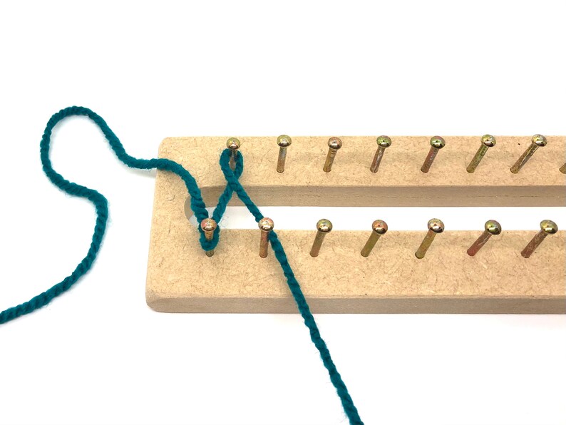 Puede incluir: Un telar de punto de madera con clavijas met&aacute;licas, parcialmente ensartado con hilo verde azulado. El telar es rectangular con dos filas de clavijas. El hilo est&aacute; enrollado alrededor de las clavijas, lo que demuestra una t&eacute;cnica de tejido. El fondo es blanco.