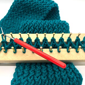 Puede incluir: Bufanda de punto color turquesa en proceso sobre un telar de madera. El telar tiene clavijas doradas y un ganchillo con mango rojo. La bufanda tiene un patr&oacute;n texturizado, y el fondo es blanco.