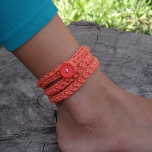Pode incluir: Uma pulseira de tornozelo de crochê laranja com fecho de botão vermelho. A pulseira envolve o tornozelo várias vezes. O fundo inclui uma perna com leggings turquesa e uma superfície de madeira.