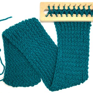 Puede incluir: Una bufanda tejida de color verde azulado con un patr&oacute;n texturizado, parcialmente terminada en un telar de madera. La bufanda est&aacute; hecha de hilo grueso y se encuentra sobre una superficie blanca, mostrando el trabajo detallado de puntadas y las clavijas del telar.