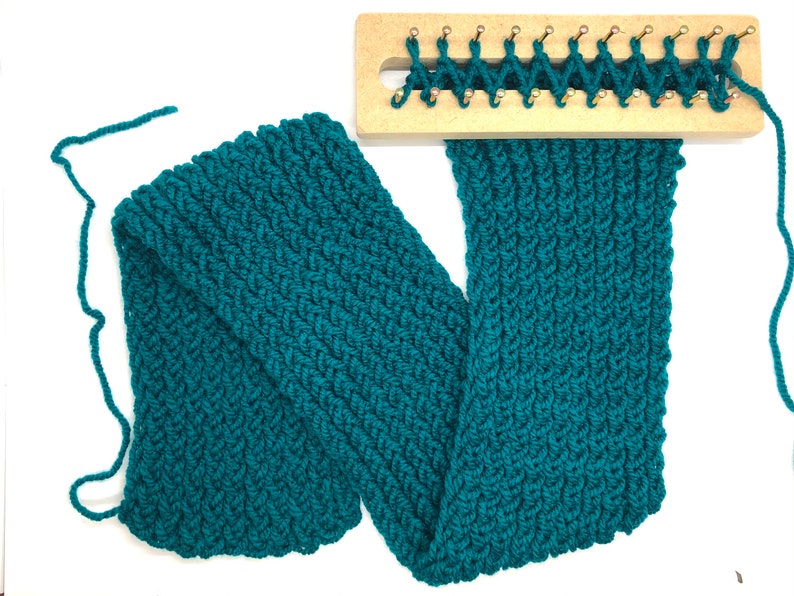 Puede incluir: Una bufanda tejida de color verde azulado con un patr&oacute;n texturizado, parcialmente terminada en un telar de madera. La bufanda est&aacute; hecha de hilo grueso y se encuentra sobre una superficie blanca, mostrando el trabajo detallado de puntadas y las clavijas del telar.