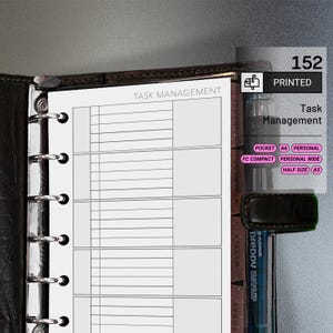 May include: A black leather-bound planner open to a task management page. The page is white with horizontal lines and the words "TASK MANAGEMENT" at the top. A clear plastic pen holder is visible, along with the text "PRINTED" and "Task Management".