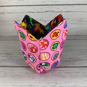 May include: A pink fabric cup with a colorful pattern of Girl Scout badges. The cup is folded into a cone shape.