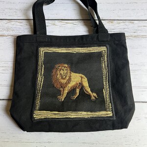 May include: Black tote bag with a rectangular embroidered design featuring a golden lion. The lion is depicted in a regal pose within a gold-colored frame. The bag has black handles and is made of a sturdy fabric.