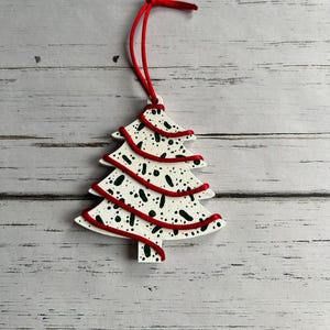 May include: A white Christmas tree ornament with red trim and a red string for hanging. The tree is decorated with black and green speckles. The ornament is on a light gray wooden surface.
