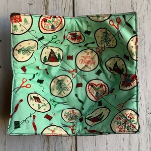 May include: A square, quilted bowl cozy in a light teal fabric. The fabric is patterned with cream-colored circles containing Christmas-themed embroidery designs, scissors, and spools of thread. The words "Merry Christmas" are visible on one circle.