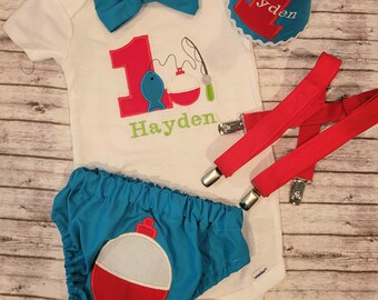 May include: A white onesie with a red number one and a fishing bobber design. The onesie has a blue bow tie and the name Hayden is embroidered on it. A blue and white hat with the number one and the name Hayden is included. Red suspenders and blue shorts with a fishing bobber design are also included.