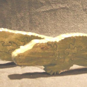 May include: Two light green wooden alligator figures, each constructed as a jigsaw puzzle. The figures feature textured surfaces and visible interlocking puzzle piece seams that form the shape of the reptiles, including heads, bodies, and tails.