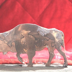 May include: A wooden puzzle shaped like a bison, featuring interlocking pieces that form the animal's silhouette. The wood has a dark brown finish with visible grain and carved texture details on the head, mane, and tail areas of the bison figure.