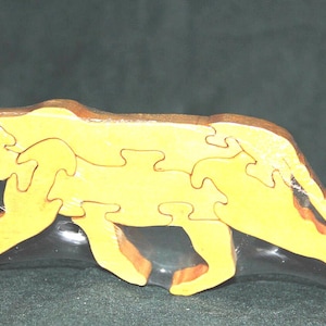 May include: A light yellow wooden puzzle shaped like a large cat, such as a panther or lion, composed of several interlocking pieces. The entire puzzle is encased in a clear, thin plastic shrink-wrap film that follows the animal's outline.