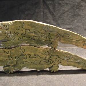 May include: Two stacked wooden alligator puzzles painted in a muted olive green color. Each puzzle is segmented into several interlocking pieces with visible dark outlines defining the shape of the alligator, including the head, body, and tail.