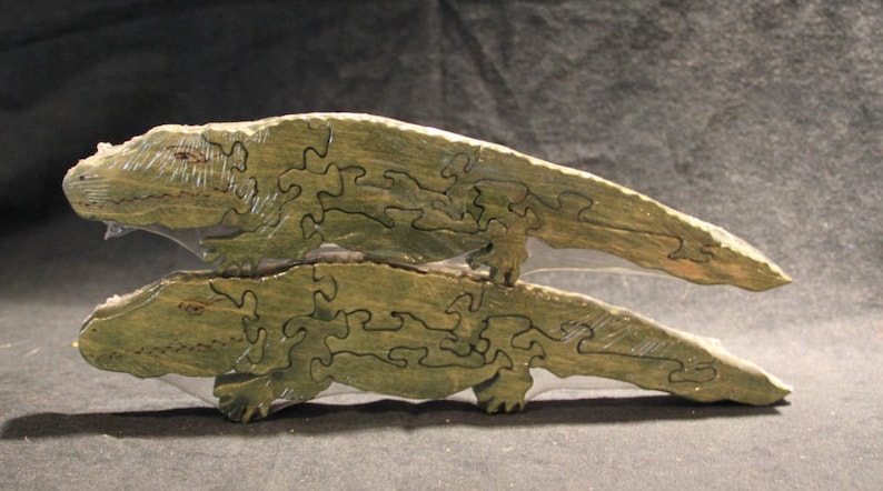 May include: Two stacked wooden alligator puzzles painted in a muted olive green color. Each puzzle is segmented into several interlocking pieces with visible dark outlines defining the shape of the alligator, including the head, body, and tail.