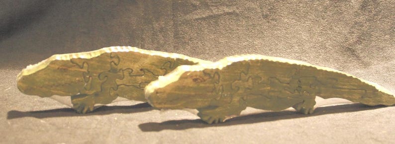 May include: Two light green wooden alligator figures, each constructed as a jigsaw puzzle. The figures feature textured surfaces and visible interlocking puzzle piece seams that form the shape of the reptiles, including heads, bodies, and tails.