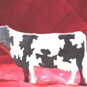 May include: A cow-shaped puzzle made of interlocking wooden pieces. The puzzle features a classic black and white spotted pattern across its body, with a distinct head, four legs, and a tail. The surface has a smooth, glossy finish.